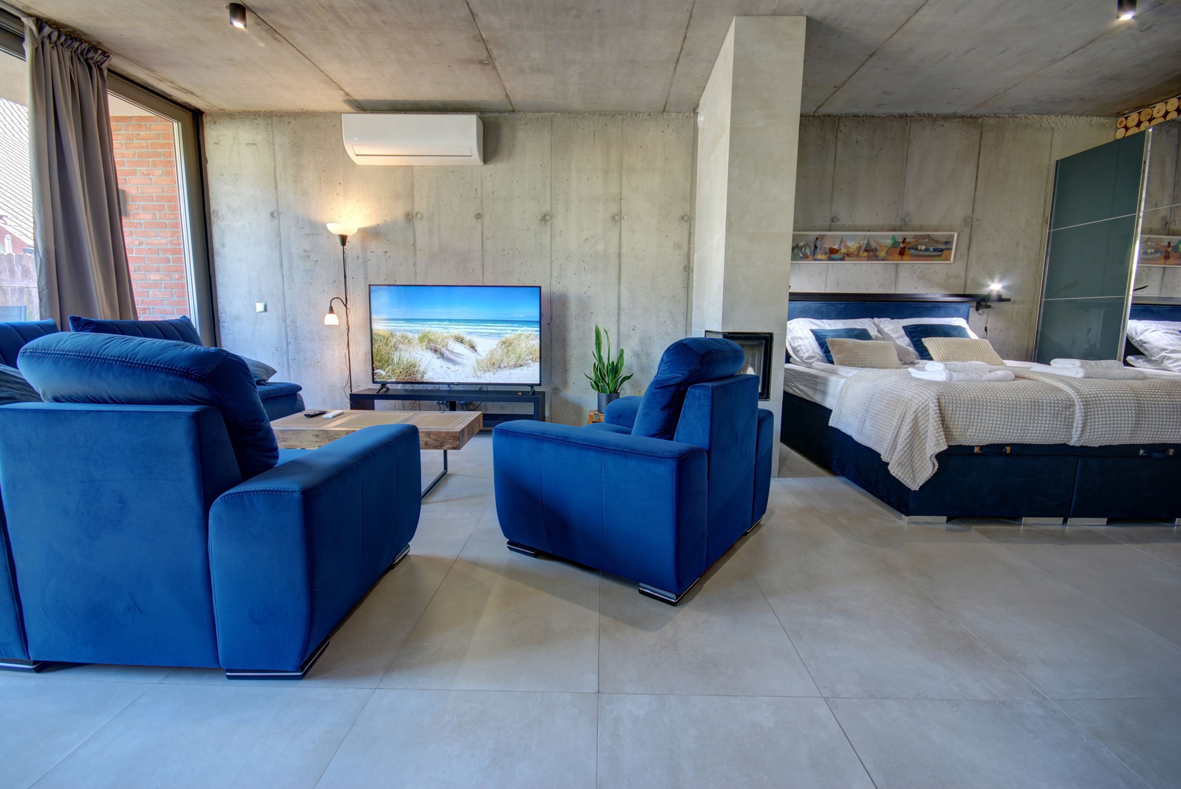 Una sala moderna con un televisor de pantalla plana grande, sofás azules, una cama con cabecera azul y una pared de ladrillo visible a través de una ventana.