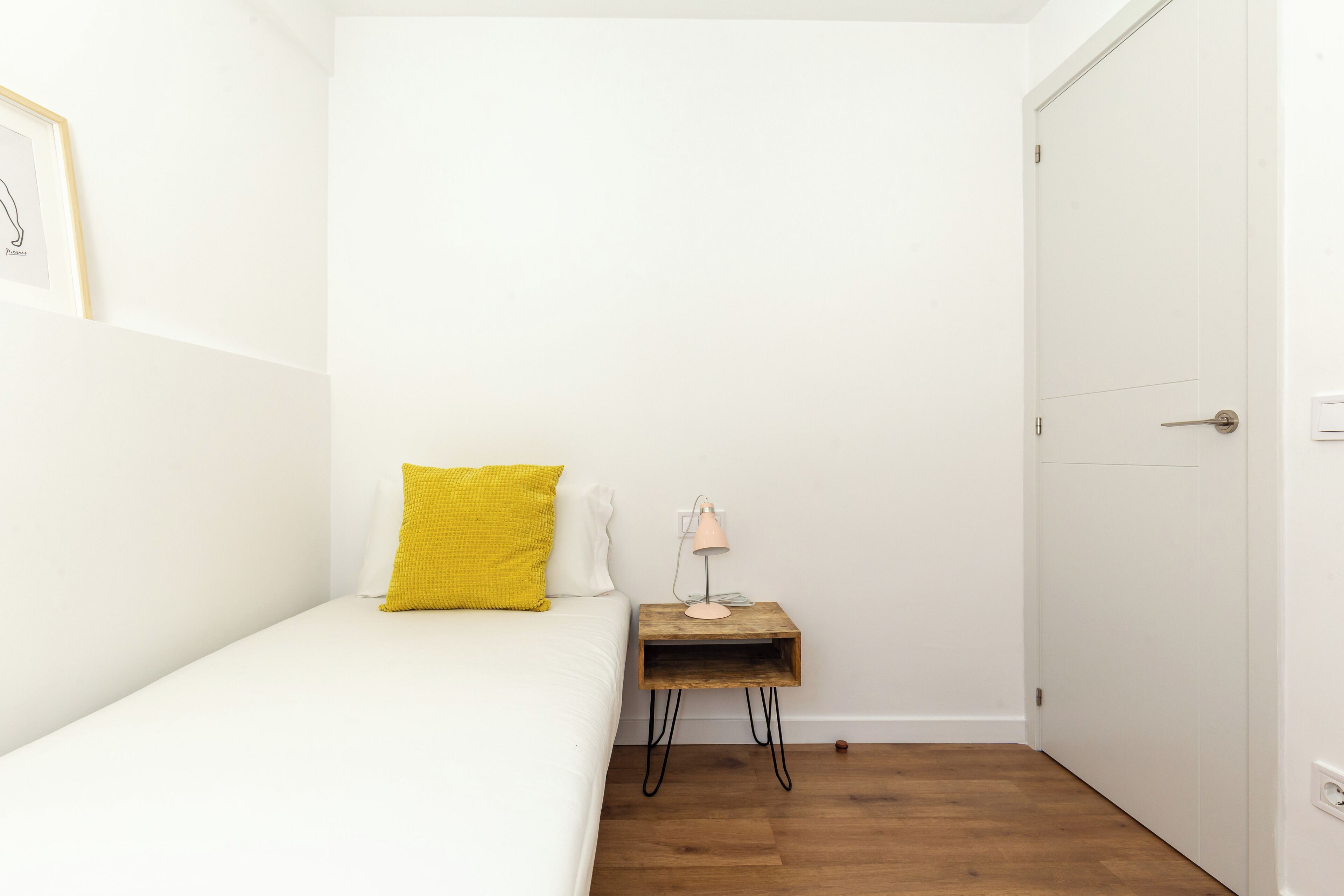 A small, white room with a single bed, a yellow pillow, a bedside table with a lamp, and a wooden floor.