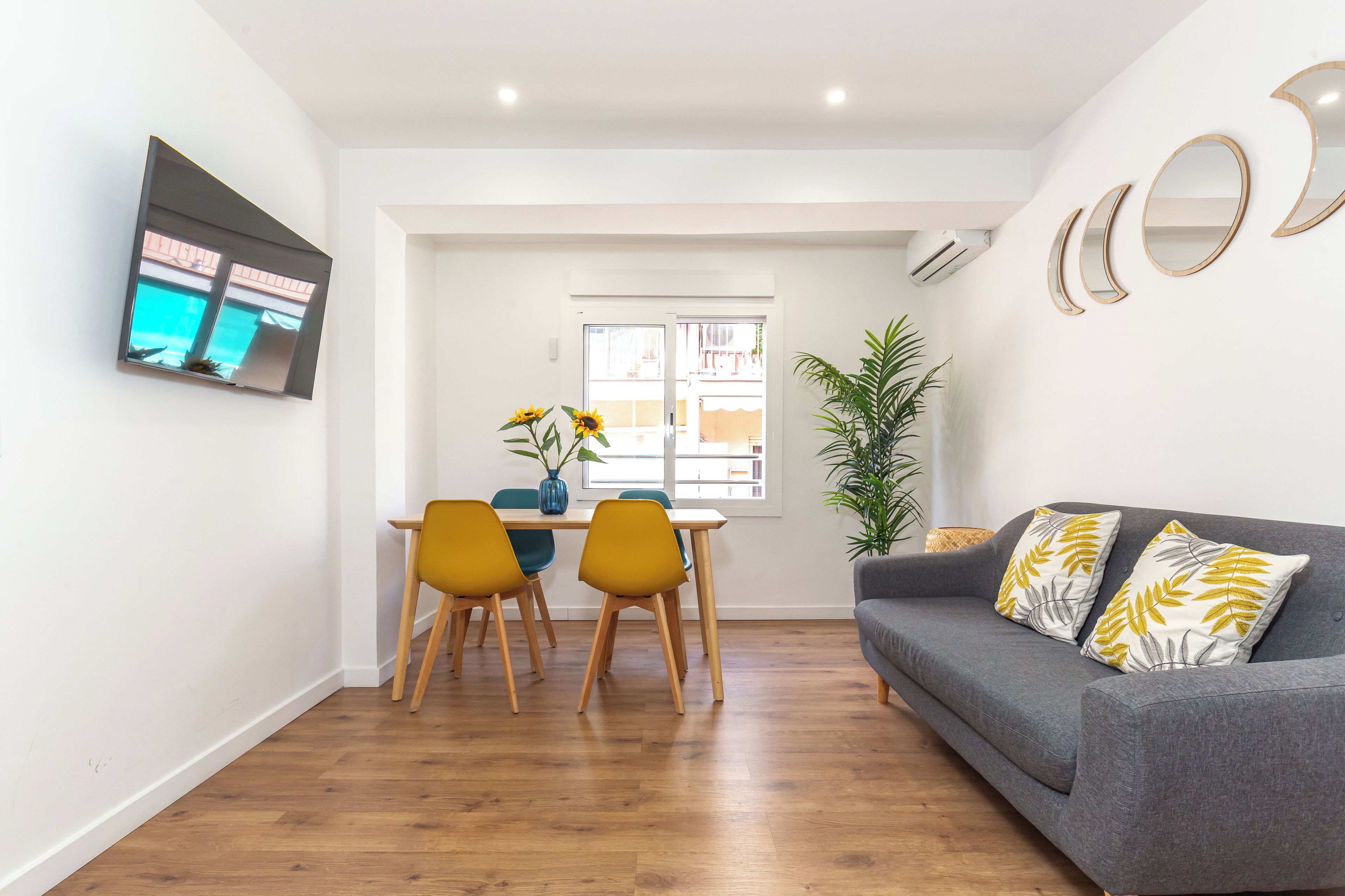 A modern living room with a grey sofa, yellow chairs, a dining table, and a TV mounted on the wall.