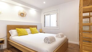 Ett modernt hotellrum med en säng i trä, vita sängkläder, en solformad väggdekoration och ett fönster med persienner.