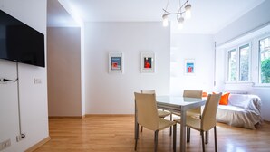 Een moderne eethoek met een houten vloer, een glazen tafel en beige stoelen. Er is een televisie aan de muur bevestigd, en er hangt kunst aan de muur.