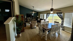 A spacious living room with a dining area, wooden floors, and a large TV. - Beach Retreat: Walk to Beach & Pool, Sleeps 12, Pet Friendly (Port Aransas)