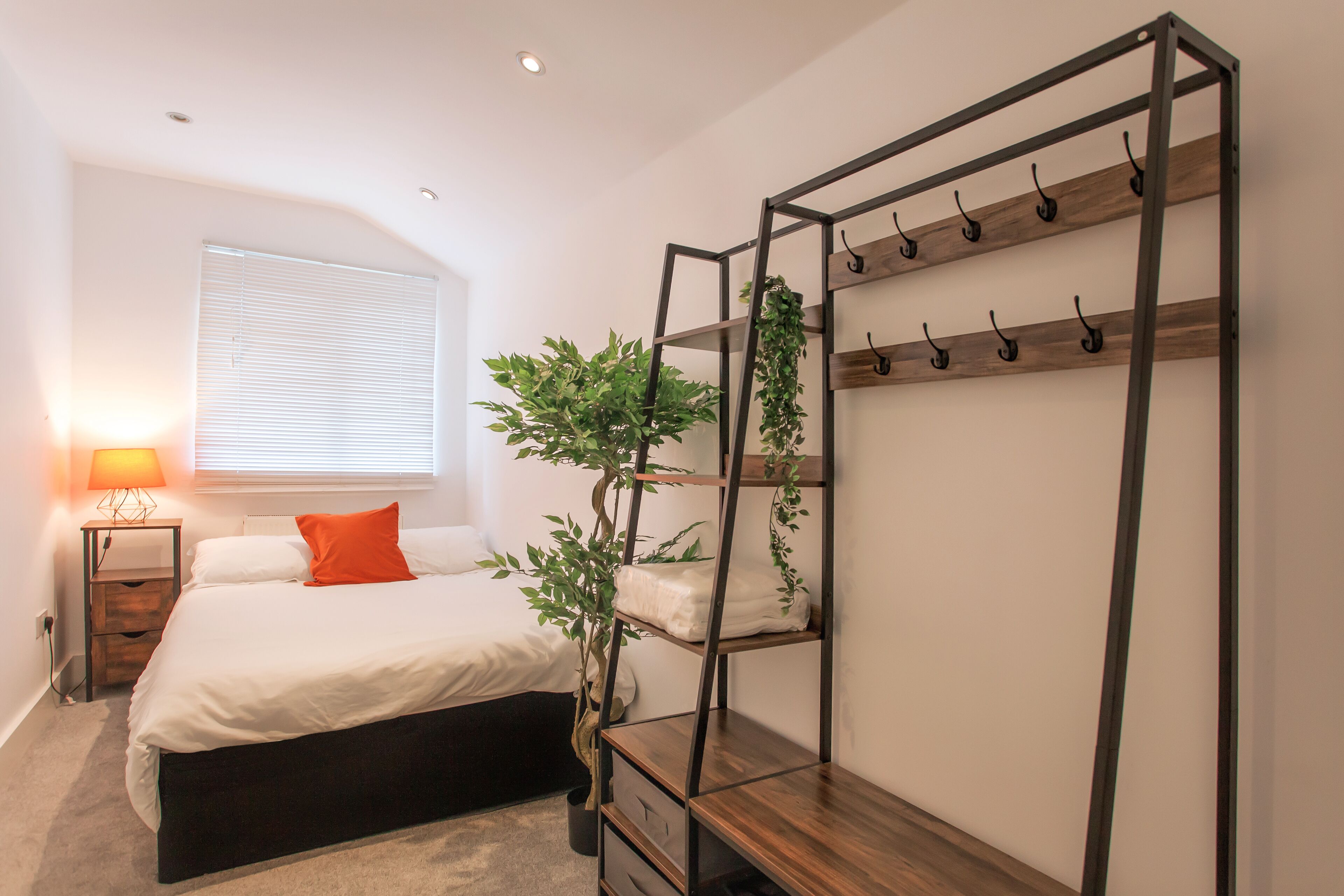 A bedroom with a bed, a bedside table, a lamp, a window with blinds, and a plant.