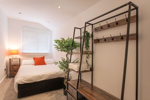 A bedroom with a bed, a bedside table, a lamp, a window with blinds, and a plant. - 3 bed House Camberwell With Private Garden (London)