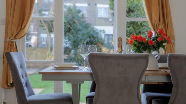 A dining area with a table set for two, a vase of red flowers, and a bottle of champagne. - 3 bed House Camberwell With Private Garden (London)