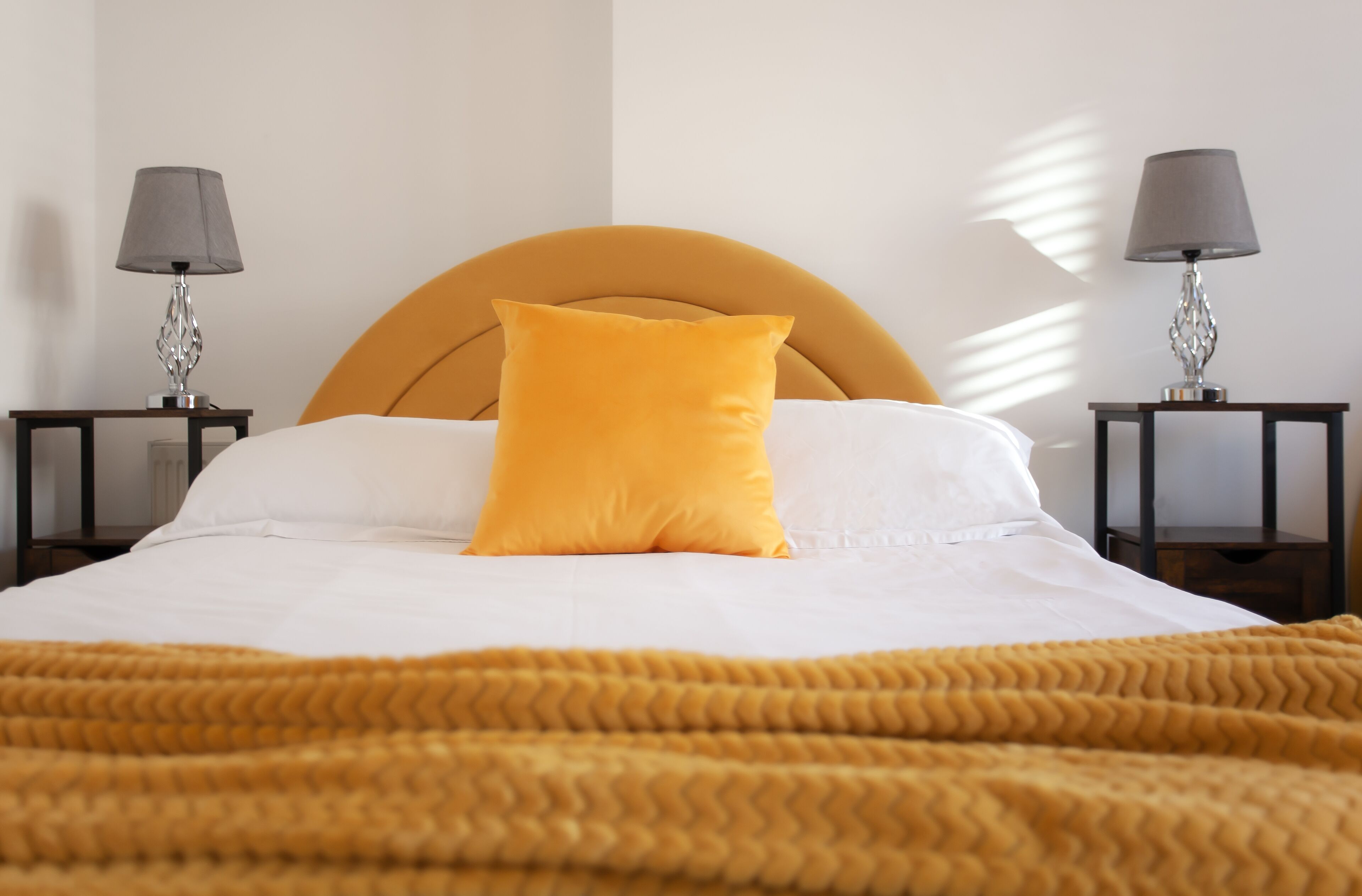 Una cama con cabecera color mostaza, un solo cojín a juego en color mostaza y una manta a juego. Hay dos mesitas de noche con lámparas a cada lado de la cama.