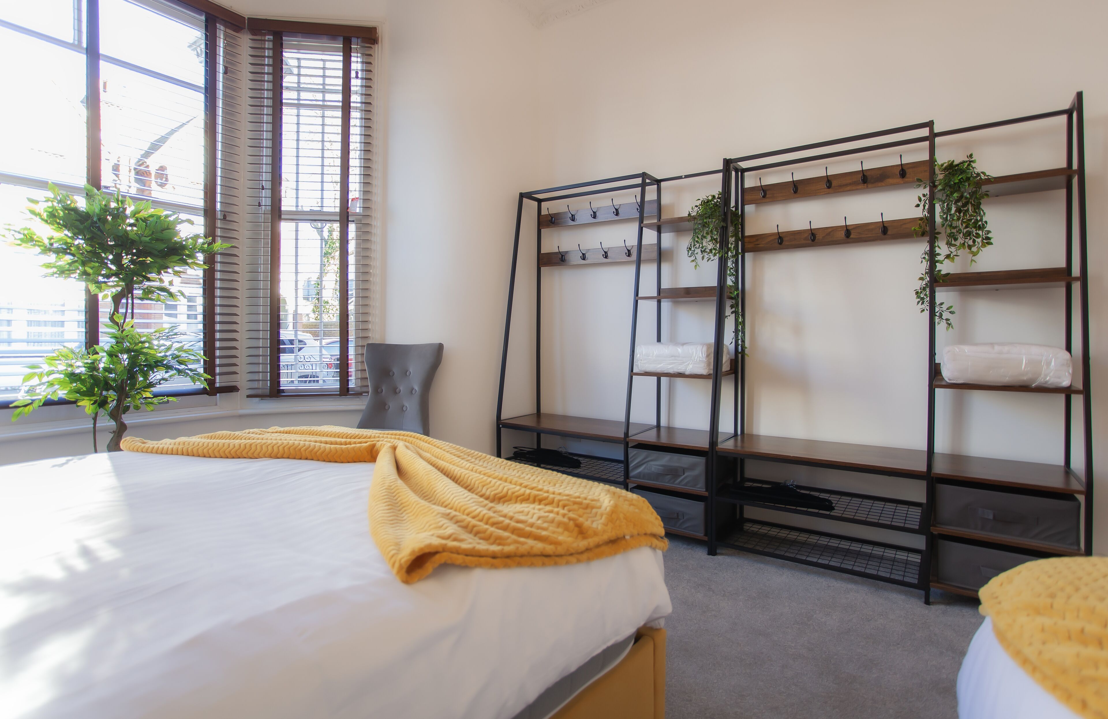 A bedroom with a bed, a window with blinds, a plant, and a metal shelf unit with hooks.