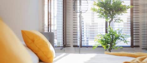 A hotel room with a bed, a window with blinds, a small plant, and a yellow pillow.