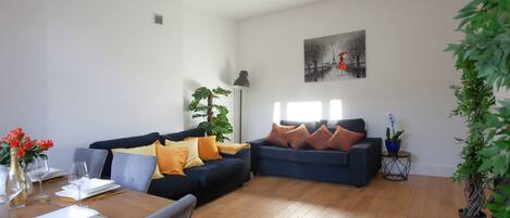 A modern living room with a dark blue sofa, orange cushions, and a dining table set for two.
