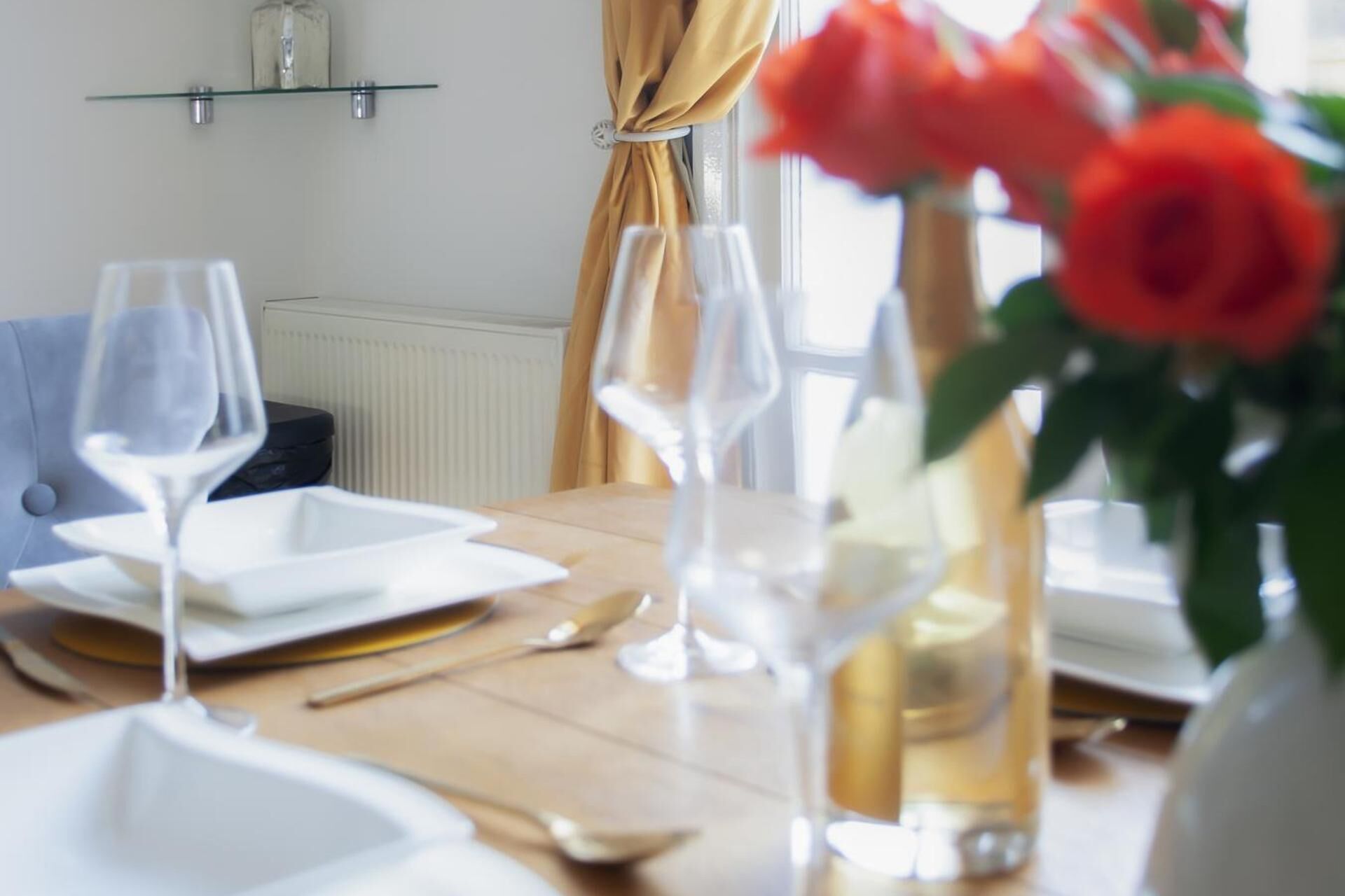 Een eettafel gedekt met witte borden, wijnglazen en bestek, met een vaas rode bloemen op de achtergrond.