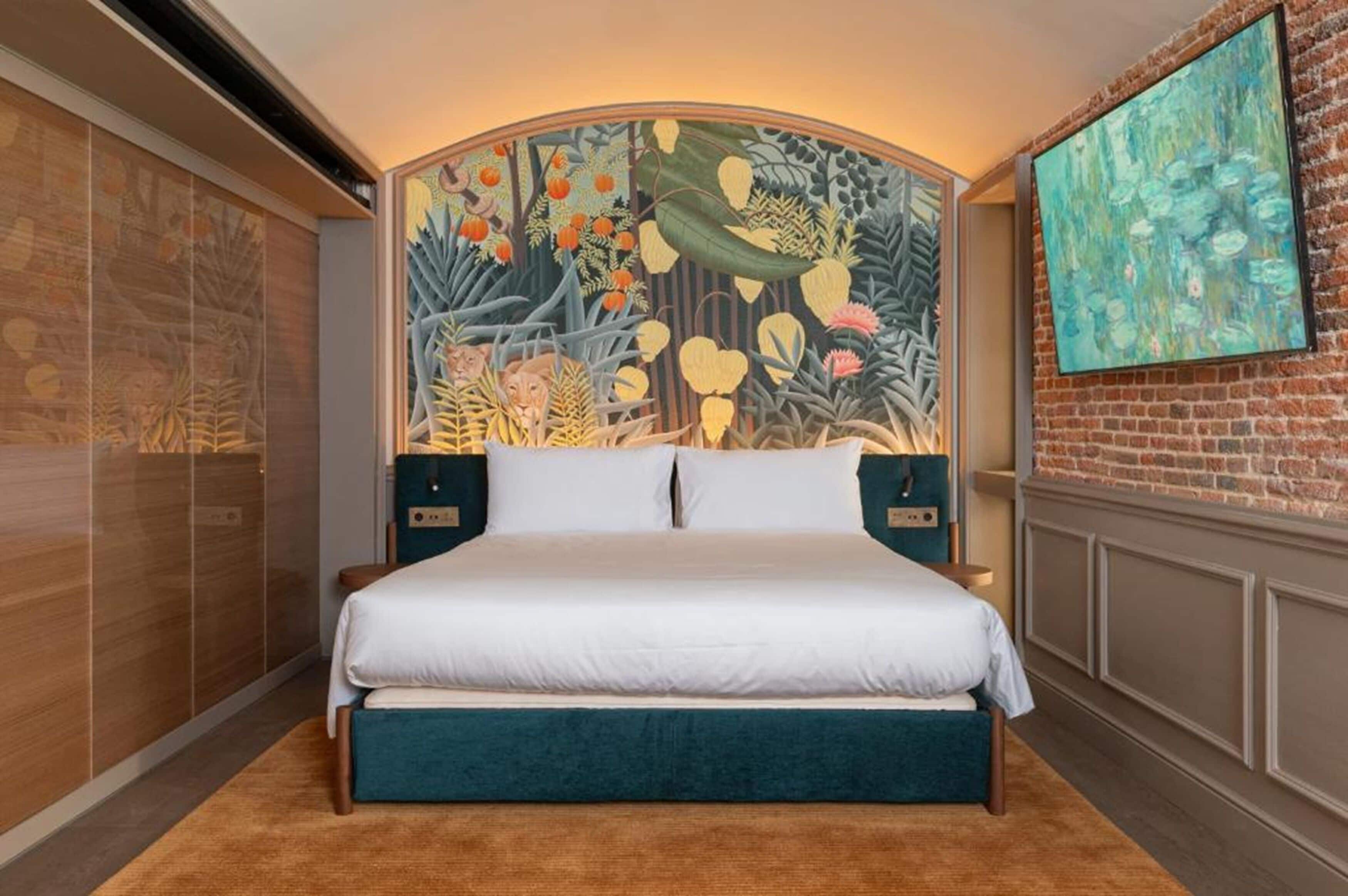 A bedroom with a large bed, a headboard with a mural, a brick wall, and a painting on the wall.