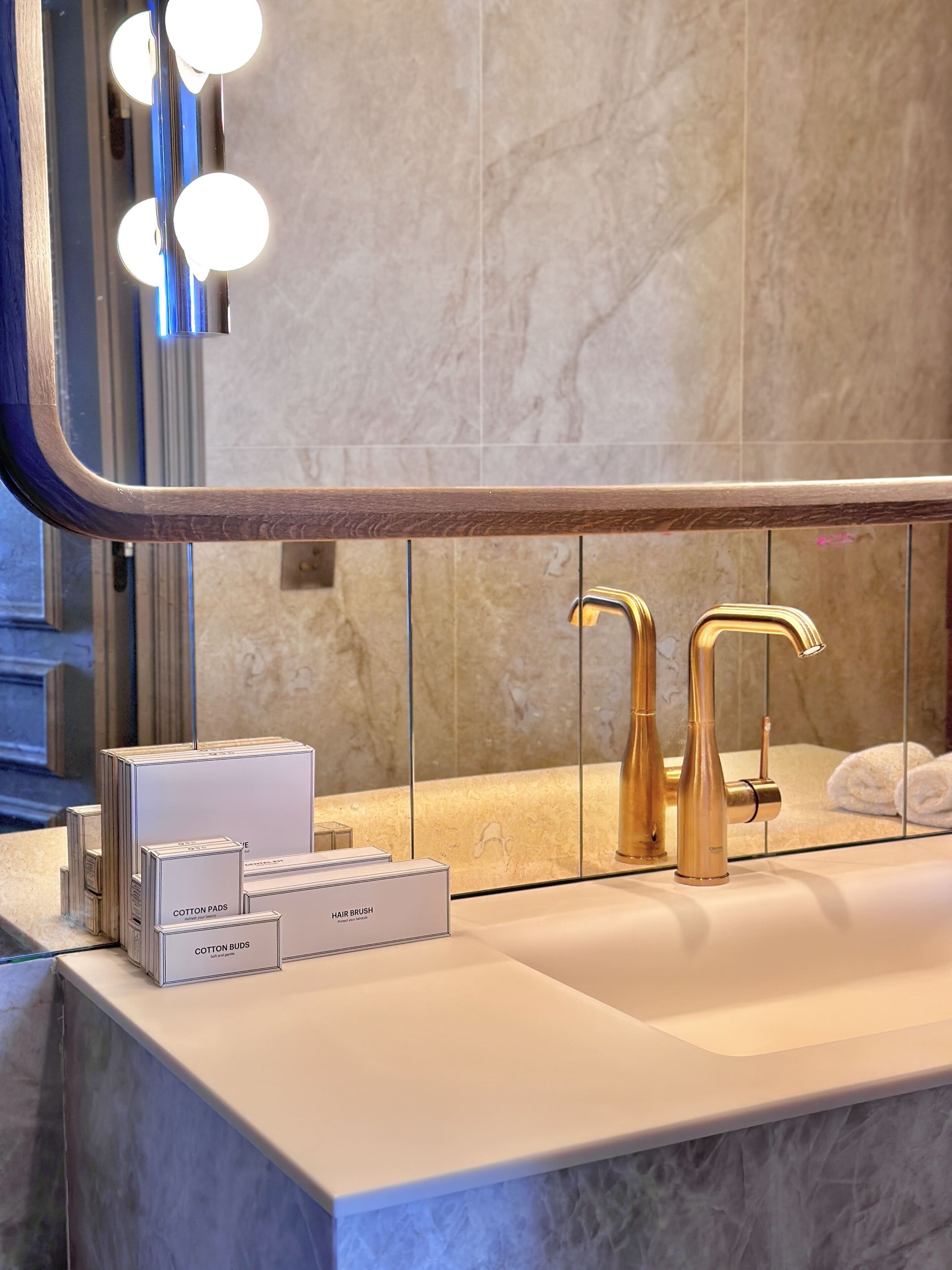 Un baño con encimera de mármol, grifo dorado y un espejo que refleja una pared azul y luces.