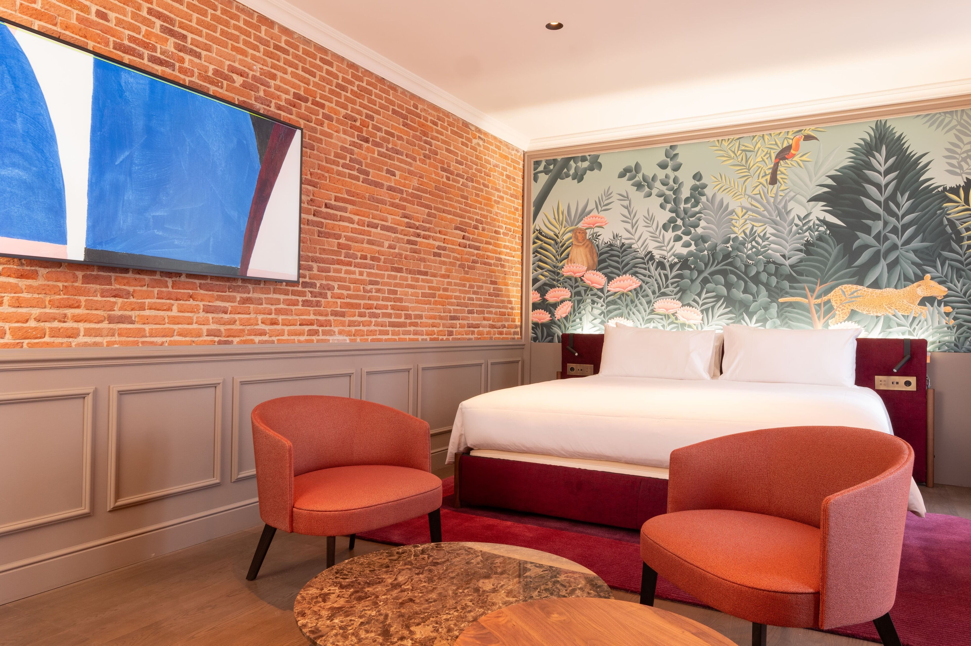 A hotel room with a brick wall, a large painting, a bed with red bedding, two orange chairs, and a round table.