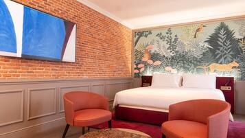 A hotel room with a brick wall, a large painting, a bed with red bedding, two orange chairs, and a round table.