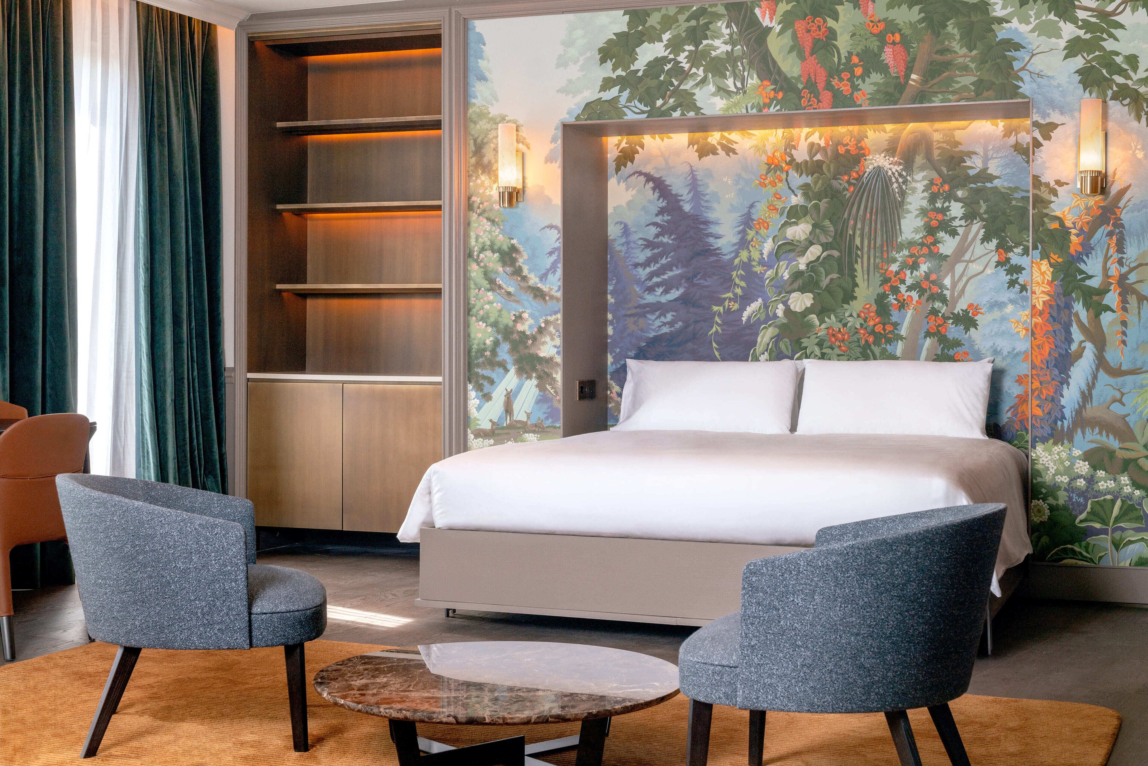 A modern hotel room with a large bed, two armchairs, a small round table, and a bookshelf.