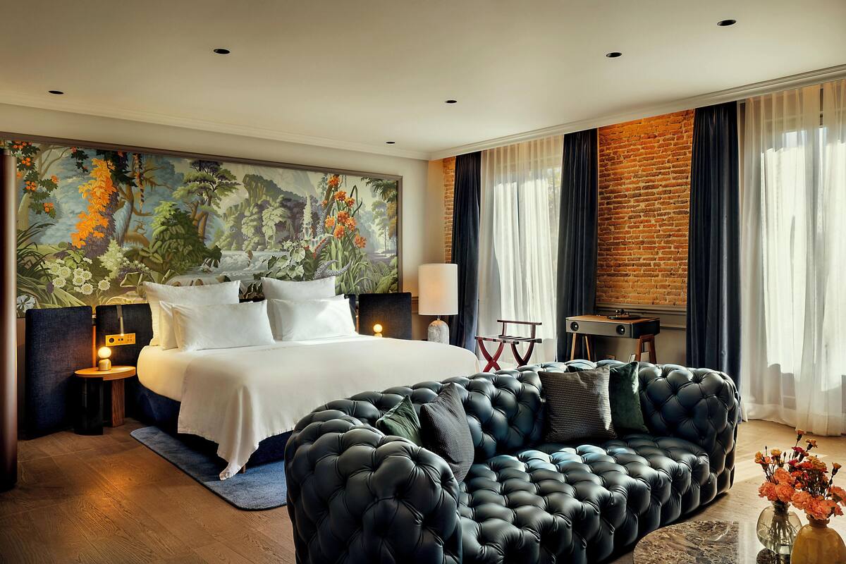 A modern hotel room with a large bed, a black tufted sofa, a mural on the wall, and a brick accent wall.
