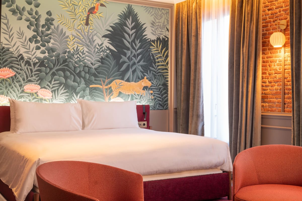 A hotel room with a bed, two chairs, a mural, a brick wall, and a window with curtains.