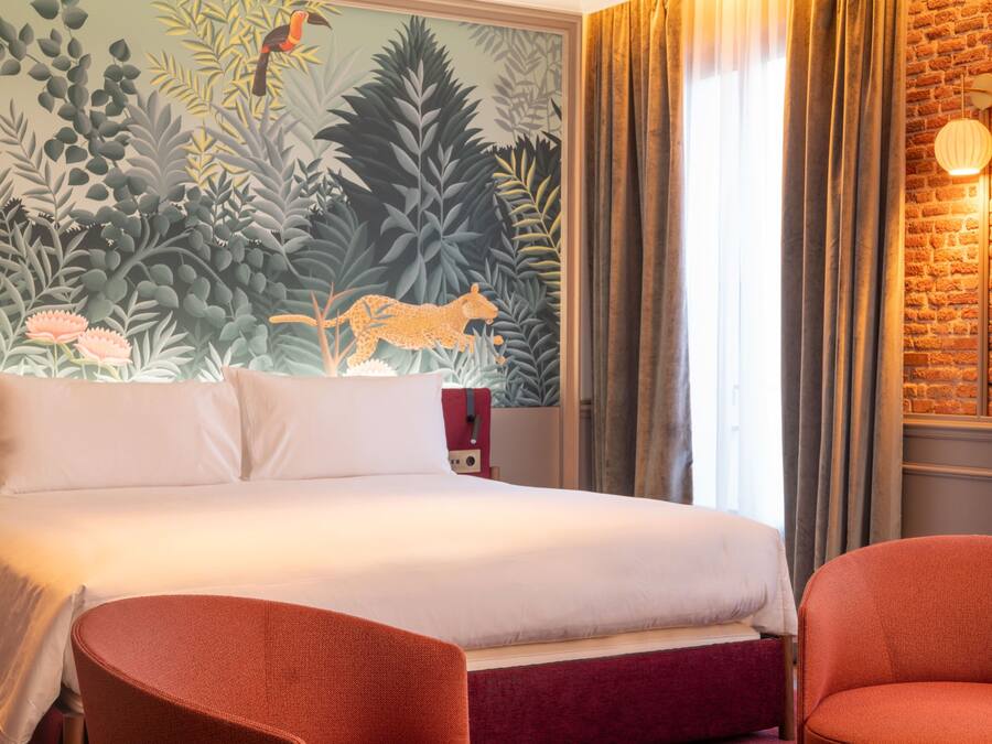 Habitació d'hotel amb un llit, dues cadires, un mural, una paret de maó i una finestra amb cortines.