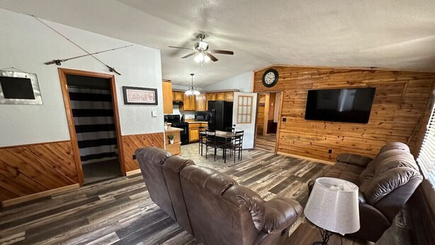 Ein gemütliches Wohnzimmer mit einer braunen Ledersofa, einem kleinen Tisch mit einer Lampe und Holzboden. Im Hintergrund ist eine Küche mit Holzschränken und eine Essecke mit Tisch und Stühlen zu sehen.