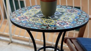 Ein kleiner runder Tisch mit einer farbenfrohen Kachelplatte, ein Blumentopf mit rosa und weißen Blüten, und ein Metallstuhl mit blauer Sitzkissen.