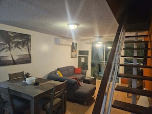 A living room with a sofa, a dining table with chairs, a ceiling fan, and a staircase.