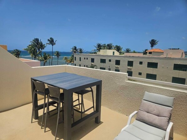 A rooftop terrace with a table and chairs, a lounge chair, and a view of the ocean and palm trees.