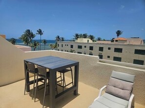 Une terrasse sur le toit, avec une table et des chaises, un fauteuil de relaxation et une vue sur l’océan et les palmiers.