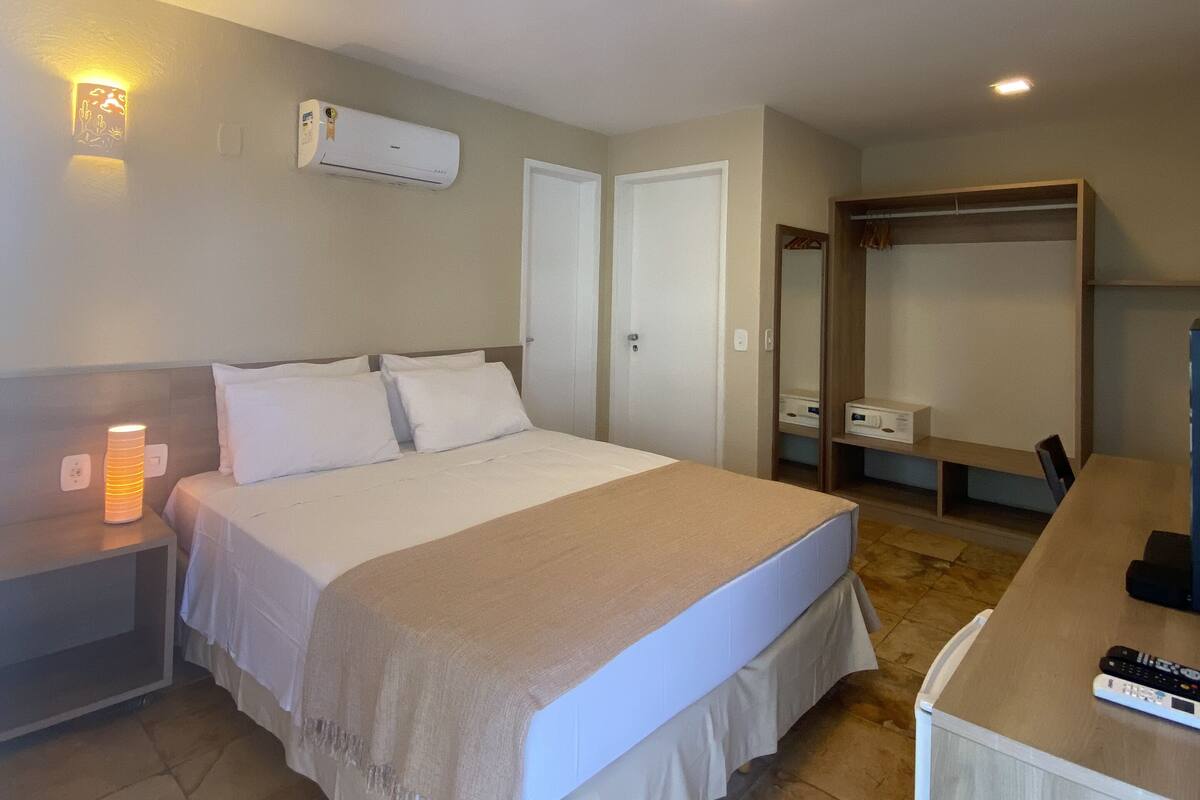 Ein Hotelzimmer mit Bett, Nachttisch, Lampe, Klimaanlage, Schrank und Schreibtisch mit Stuhl.