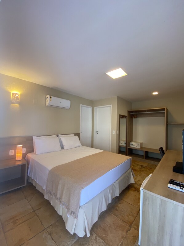 Habitación de hotel con cama, mesita de noche, lámpara, aire acondicionado, armario y un escritorio con silla.