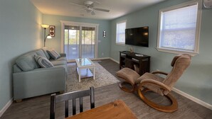 A living room with a sofa, a coffee table, a TV, a ceiling fan, and a window with blinds.