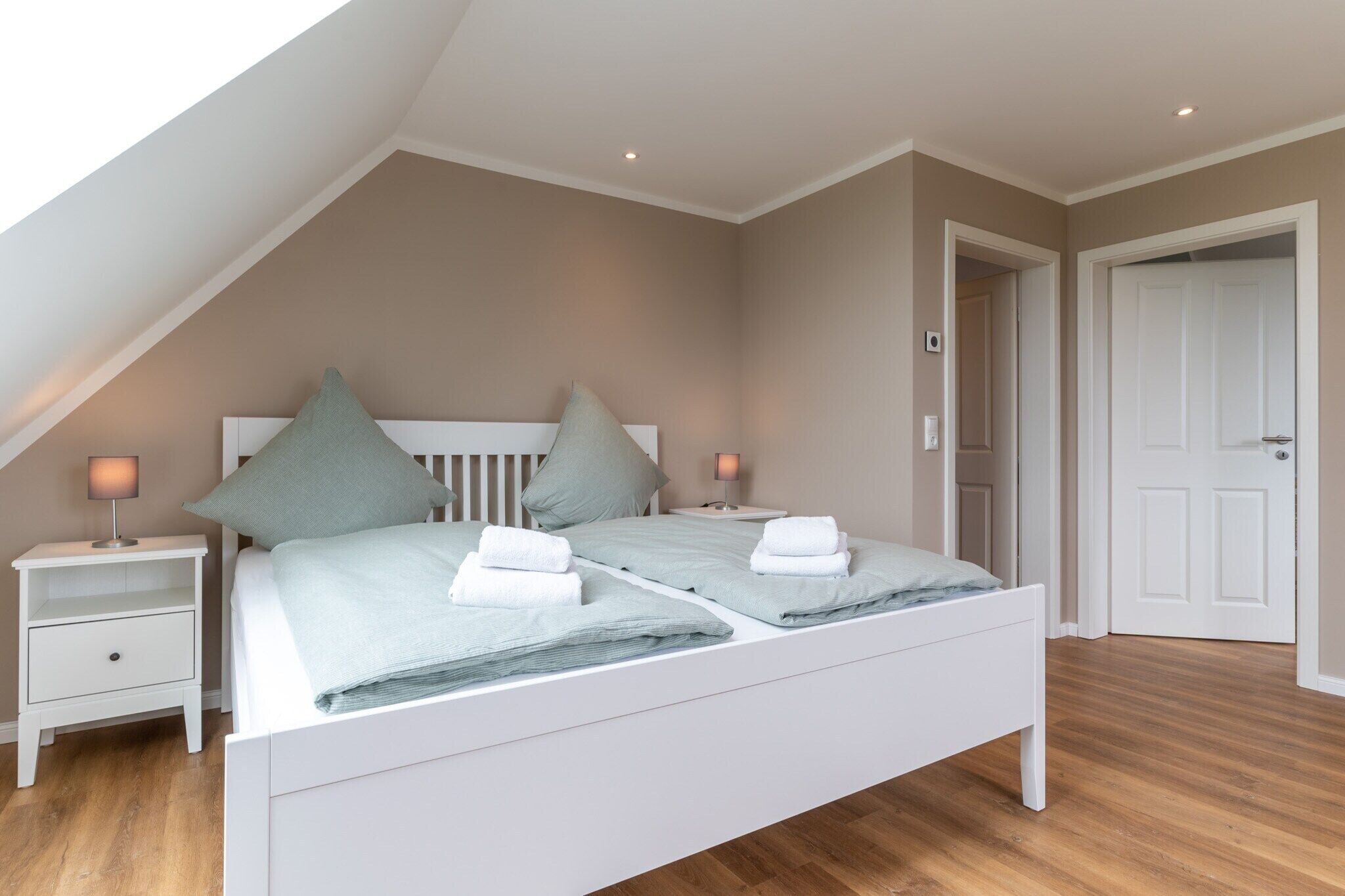 A bedroom with a sloped ceiling, a double bed with pillows and towels, bedside tables with lamps, and a door to the right.