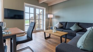 A modern living room with a wooden dining table, a sofa, and a TV mounted on the wall. - Doppelhaushälfte in Karolinenkoog (Karolinenkoog)
