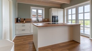A modern kitchen with a large island, white cabinetry, and a view through large windows. - Doppelhaushälfte in Karolinenkoog (Karolinenkoog)