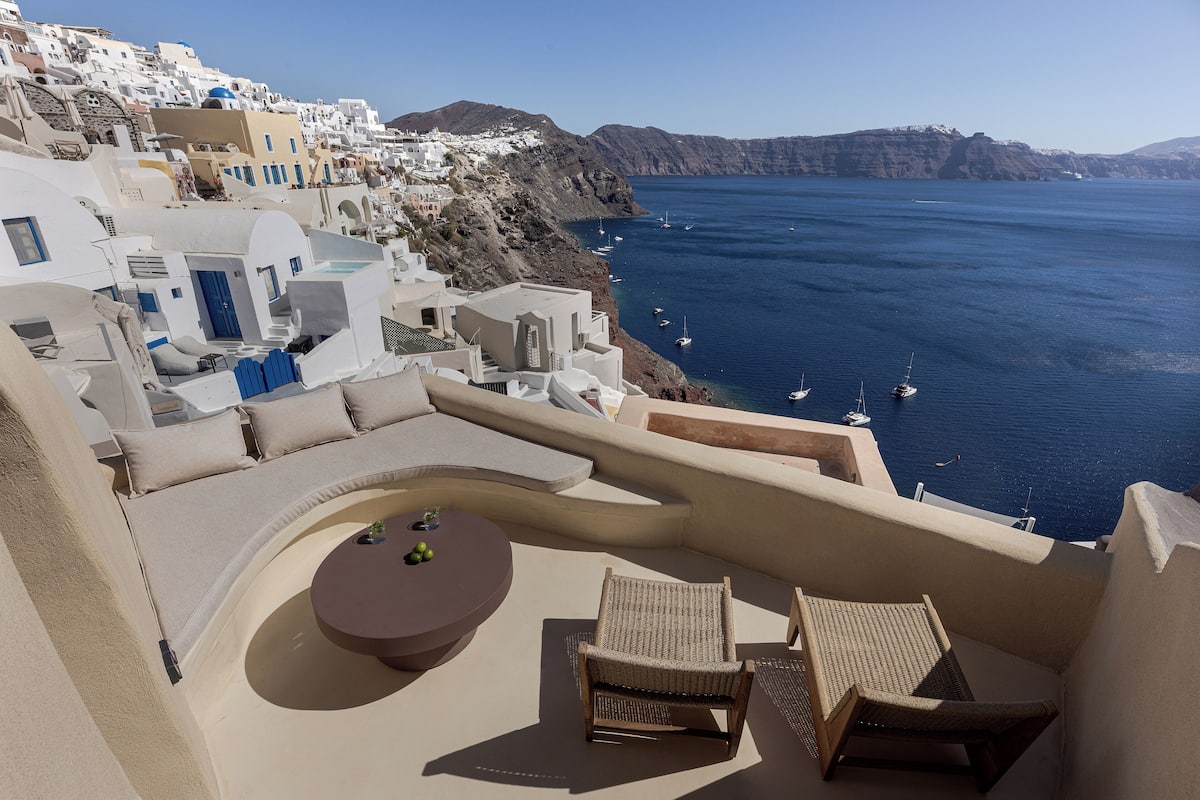 Un balcon doté d’un espace salon aux lignes courbes, d’une table ronde et de deux fauteuils en osier, offrant une vue sur une ville côtière aux bâtiments blancs et aux bateaux qui flottent sur l’eau.