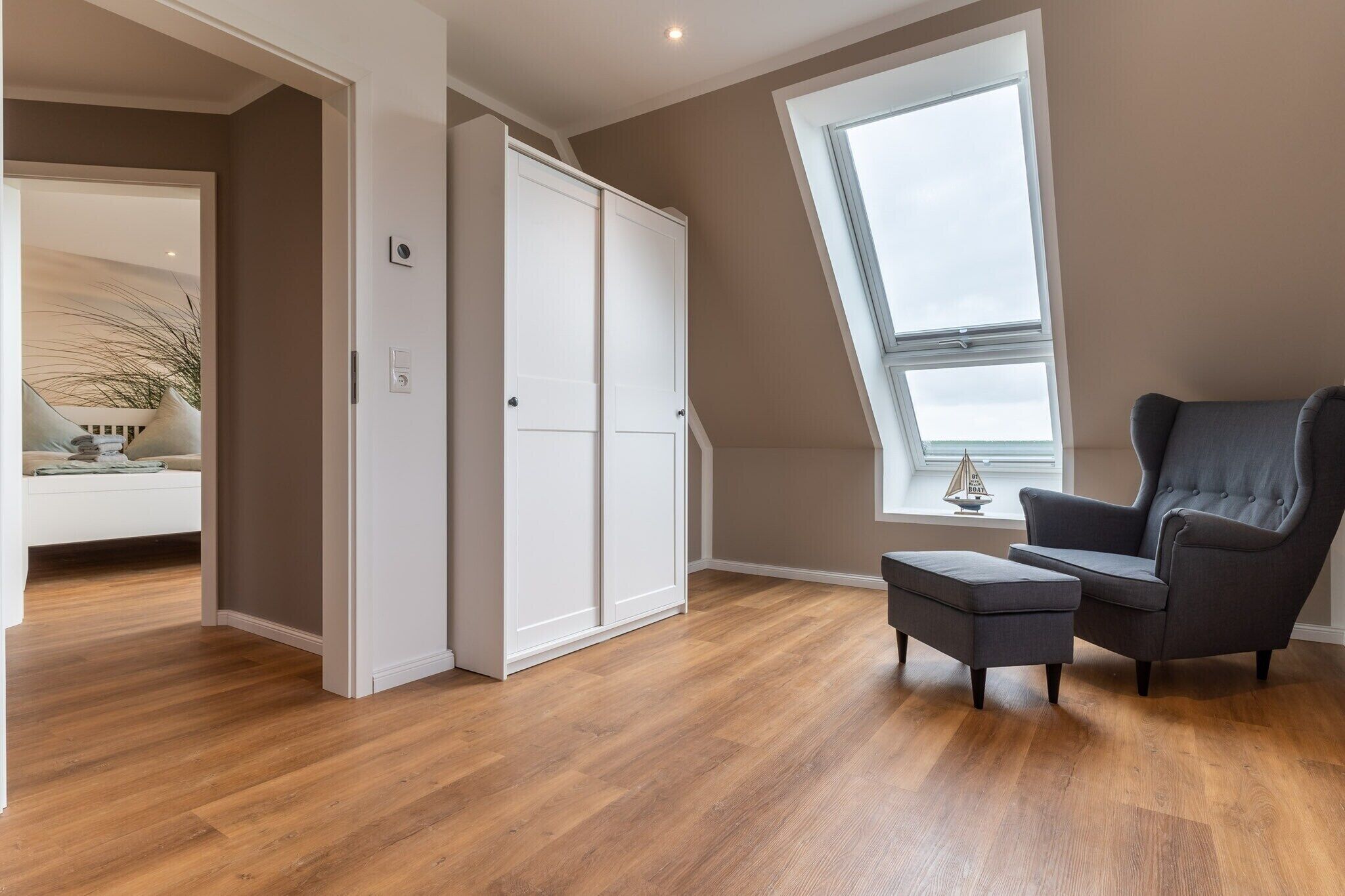 Een kamer met een groot raam, een grijze fauteuil, een witte kledingkast en een houten vloer.