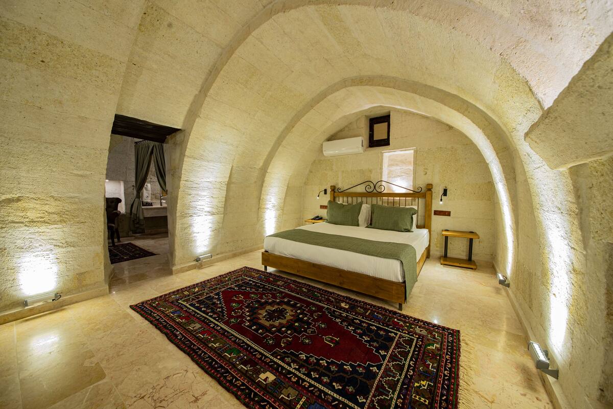 A bedroom with a large bed, a wooden headboard, a bedside table, a nightstand, a mirror, and a patterned rug.