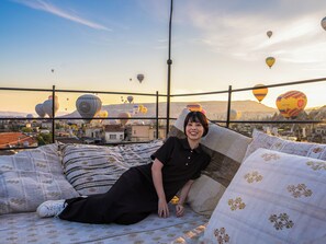 A person reclining on a bed with patterned bedding, overlooking a cityscape with hot air balloons in the distance. - Ares Cave Suites (Nevsehir)