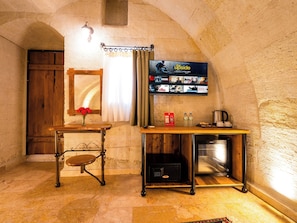 A cozy room with a stone wall, a wooden door, a table with a vase of flowers, a flat-screen TV displaying a movie, and a coffee maker. - Ares Cave Suites (Nevsehir)