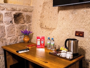 A wooden table with a remote control, a vase with purple flowers, a red box, two water bottles, a tray with snacks, a coffee maker, and a television mounted on a stone wall. - Ares Cave Suites (Nevsehir)