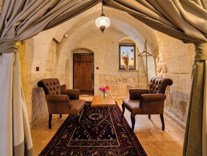 Una habitación con un techo alto en arco, una puerta de madera, dos sillones de cuero, una mesa con un tapete estampado y una lámpara colgante.