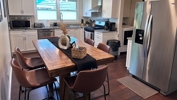 Une cuisine moderne avec des meubles blancs, un réfrigérateur en acier inoxydable, une table à manger en bois avec des chaises et une fenêtre avec des stores.