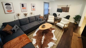 Un salon moderne avec un canapé gris foncé, des fauteuils blancs, une table basse ronde et un tapis en peau de vache. Des œuvres d’art encadrées ornent le mur, et une plante en pot est placée près de la fenêtre.