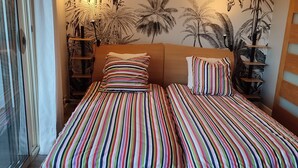 A bedroom with two beds, striped bedding, a wooden headboard, a wall mural, and a shelf with plants. - Studio Pour Cures Thermales Entièrement Refait et Équipé Pour 2 Personnes (Gréoux-les-Bains)