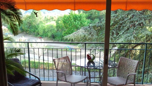 A balcony with two wicker chairs and a small table, overlooking a road and greenery. - Studio Pour Cures Thermales Entièrement Refait et Équipé Pour 2 Personnes (Gréoux-les-Bains)
