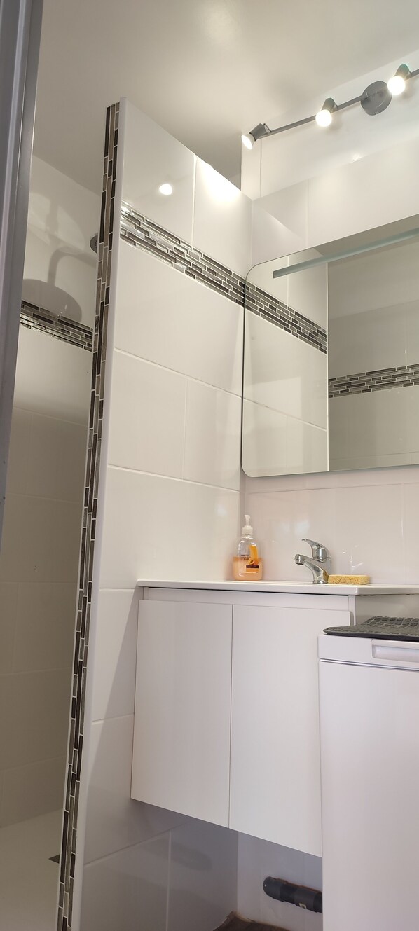 A bathroom with white tiles, a mirror, a sink with a soap dispenser, and a towel rack. - Studio Pour Cures Thermales Entièrement Refait et Équipé Pour 2 Personnes (Gréoux-les-Bains)