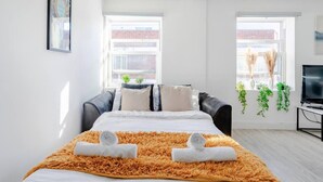 A modern bedroom with a bed, a black leather sofa, a TV, and two windows with views of a brick building. - 1-bed in Bricklane / Shoreditch - 6 Sleeps (London)
