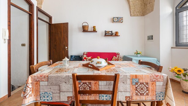 Ein Essbereich mit einem Tisch, der mit einer gemusterten Tischdecke bedeckt ist, Holzstühlen, einer roten Bank und einem an der Wand befestigten Regal mit Dekoration.