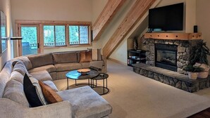 A spacious living room with a large sectional sofa, a stone fireplace, and a television mounted on the sloped ceiling. - Grand Lodge 26 at the Official Collins Lake Resort (Government Camp)