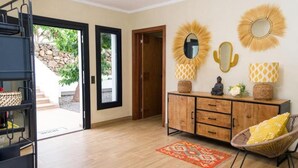 Een ruime kamer met houten vloer, een groot kastje met decoratieve spiegels, een rieten stoel en uitzicht op een stenen muur en groen buiten.