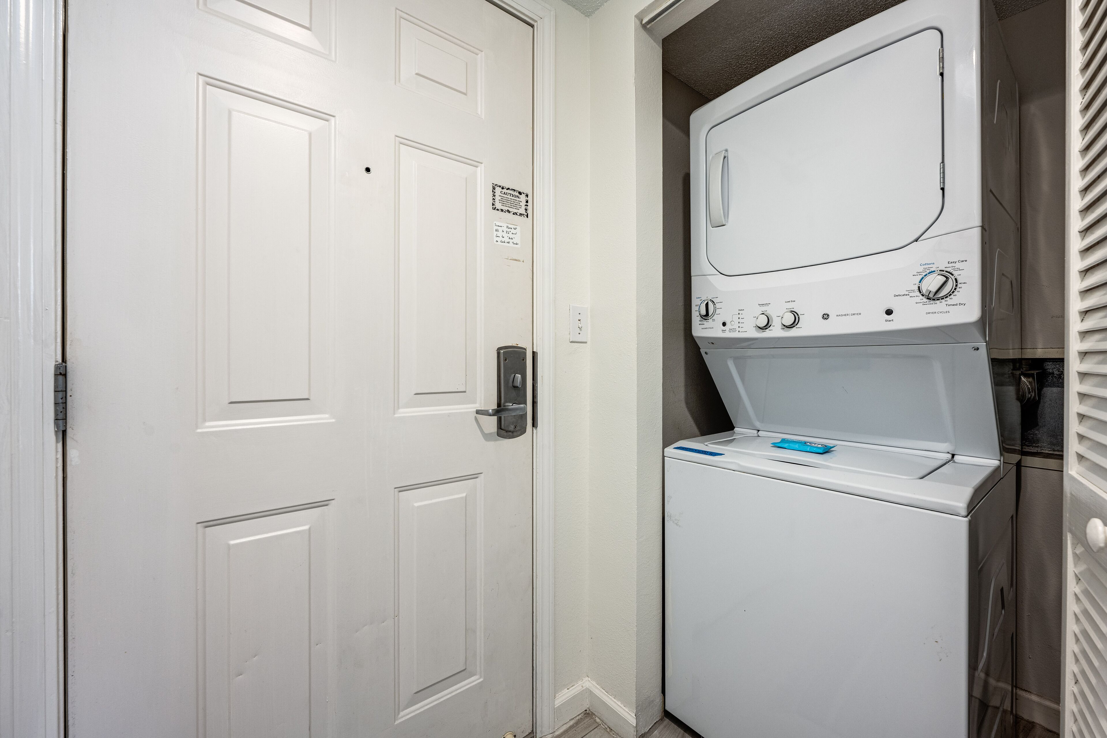 A laundry room with a front-loading washer and dryer, a door with a lock, and a small sign on the wall.